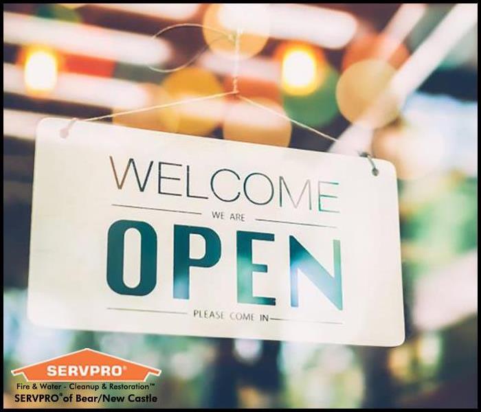 Image focused on a storefront's window where a sign reads "Welcome we are OPEN please come in"